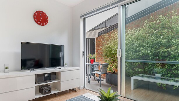 A modern living room with a flat-screen TV, a white entertainment unit, and a large sliding glass door leading to an outdoor patio with a table, chairs, and potted plants. - Hobart Cityscape (Hobart)