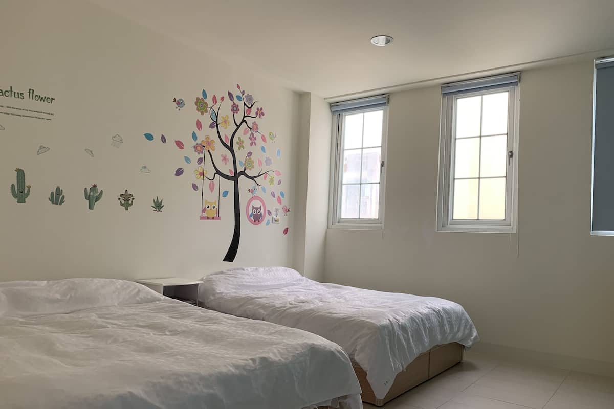 A bedroom with two beds, a wall mural of a tree with flowers and birds, and two windows.