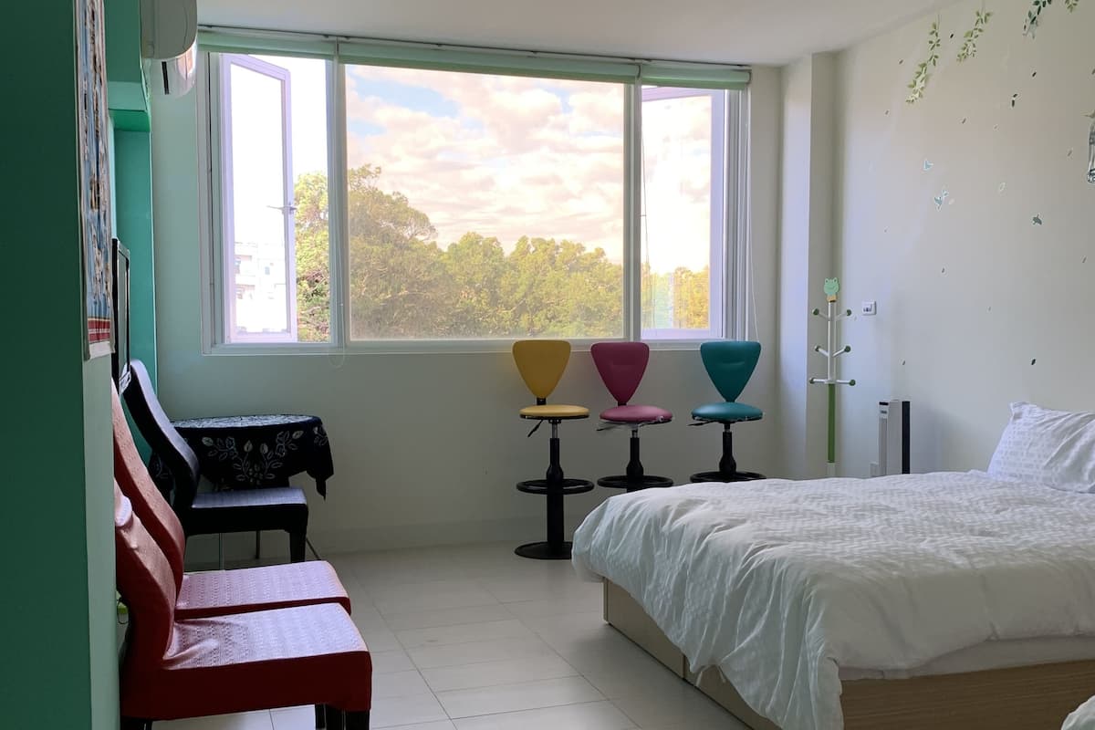 A bedroom with a bed, a window with a view of trees, a desk with a chair, and a wall-mounted coat rack.