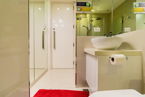 Un baño moderno con cabina de ducha de vidrio, un lavabo blanco con lavamanos y un tapete rojo en el suelo.