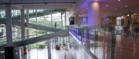 A modern shopping mall interior with a glass bridge, escalators, and a digital information board.