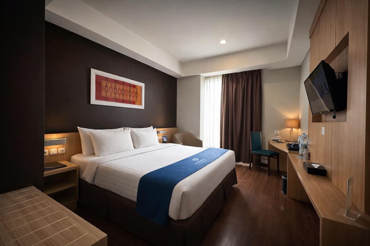 Quarto de hotel com uma cama grande, uma escrivaninha, uma cadeira, uma televisão e uma janela com cortinas.