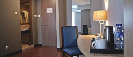 A hotel room with a wooden floor, a bed, a chair, a desk with a lamp, a coffee maker, and a water bottle.