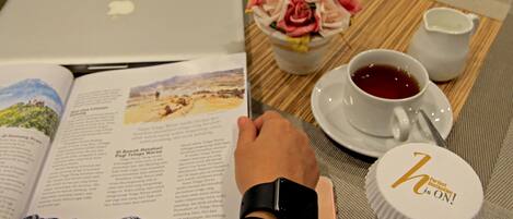 Une personne est assise à une table, un livre ouvert devant elle, un ordinateur portable à portée de main, une tasse de thé, une bouteille et une assiette de fleurs à proximité.