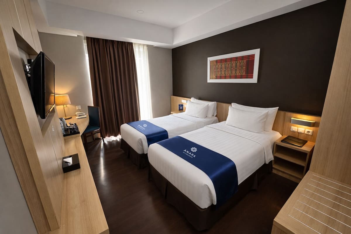 Quarto de hotel com duas camas, televisão, secretária, cadeira e janela com cortinas.