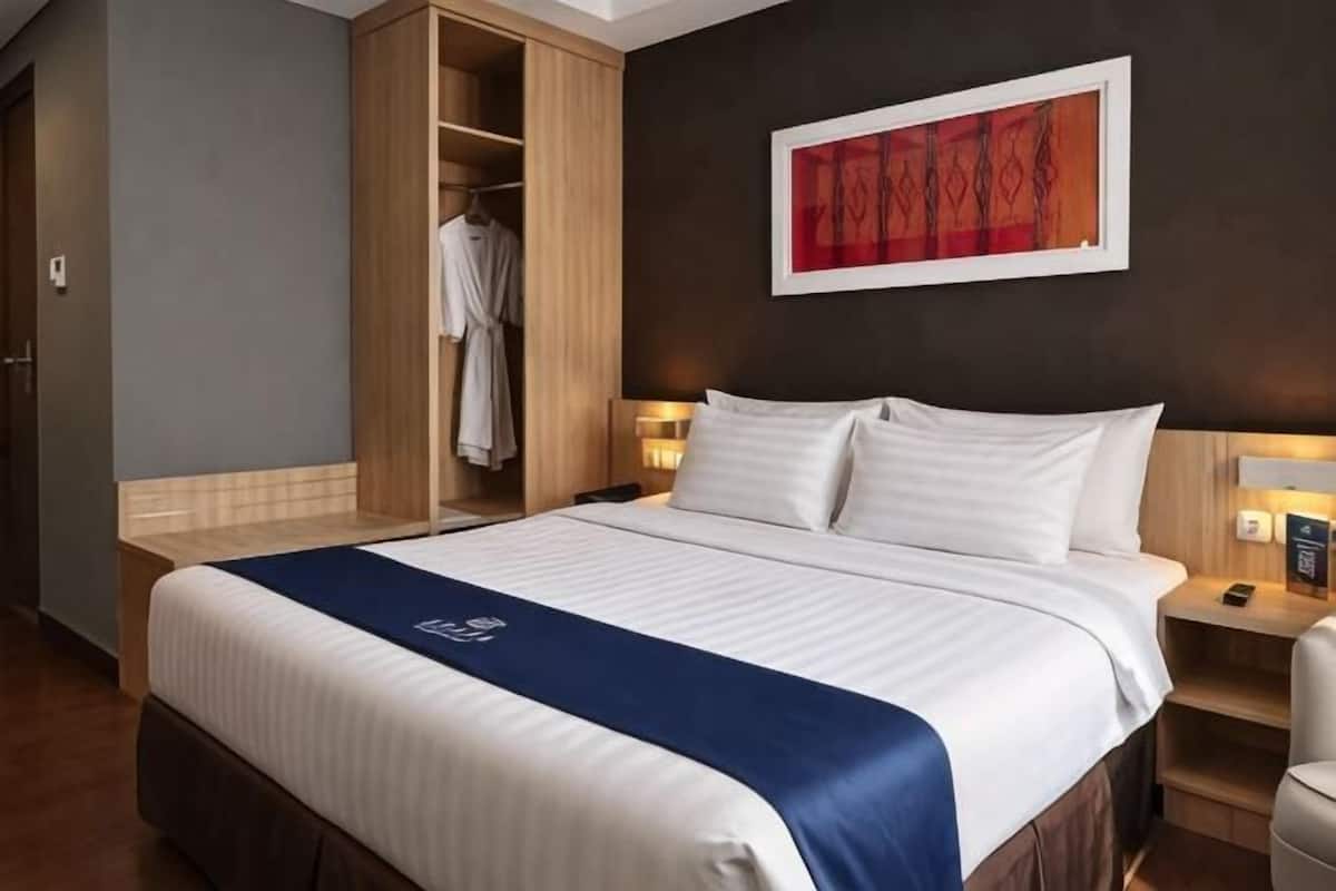 Quarto de hotel com uma cama grande, um guarda-roupa, uma mesa de cabeceira com luminária e um quadro na parede.