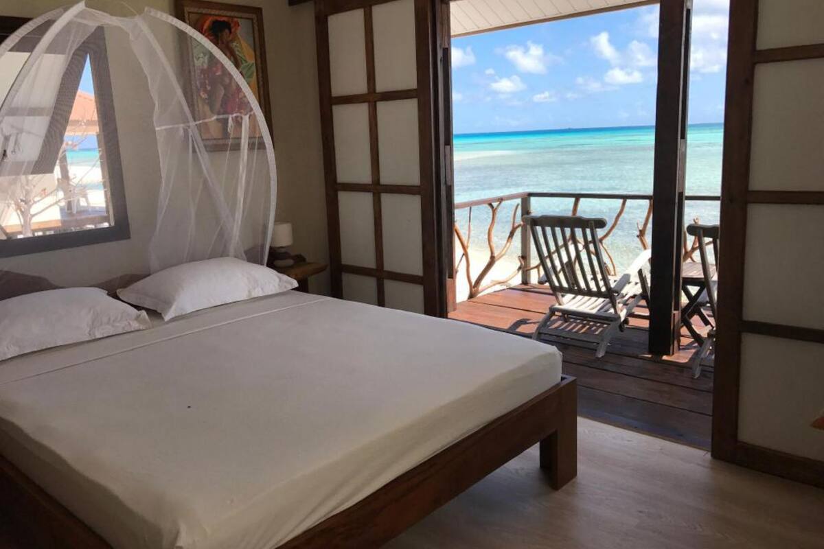 A bedroom with a bed, a window with a mosquito net, and a view of the ocean.