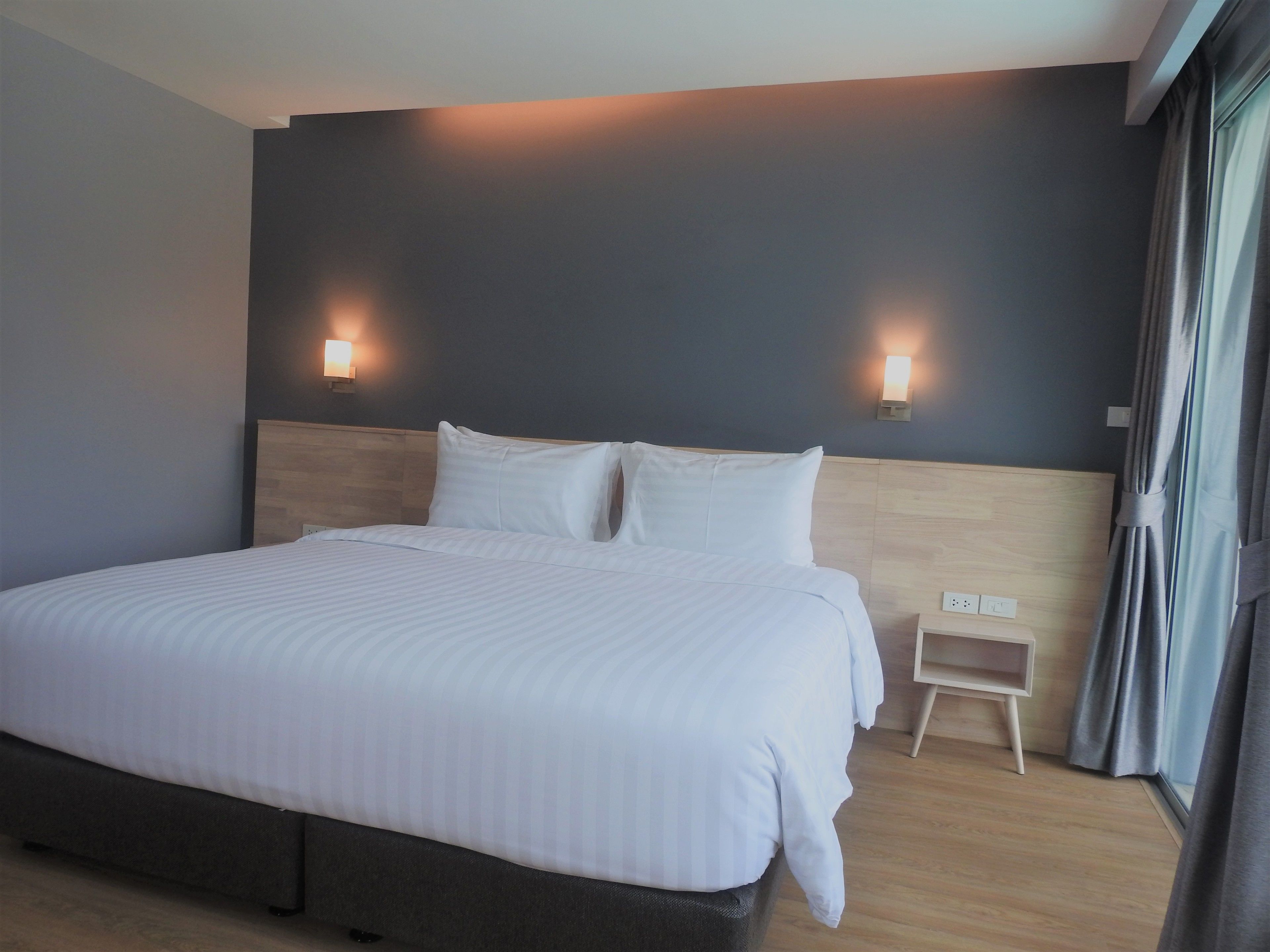 A modern hotel room with a large bed, two wall-mounted lights, a small bedside table, and a window with curtains.
