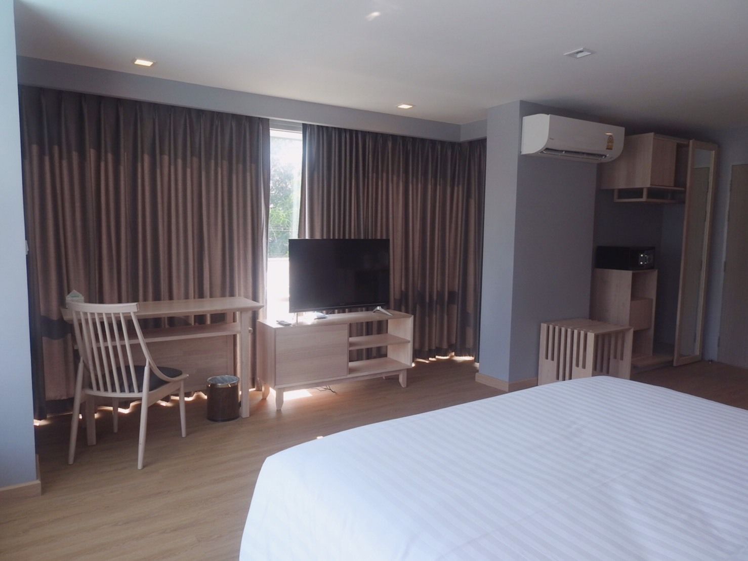 A hotel room with a bed, a TV on a wooden stand, a desk with a chair, and an air conditioning unit.