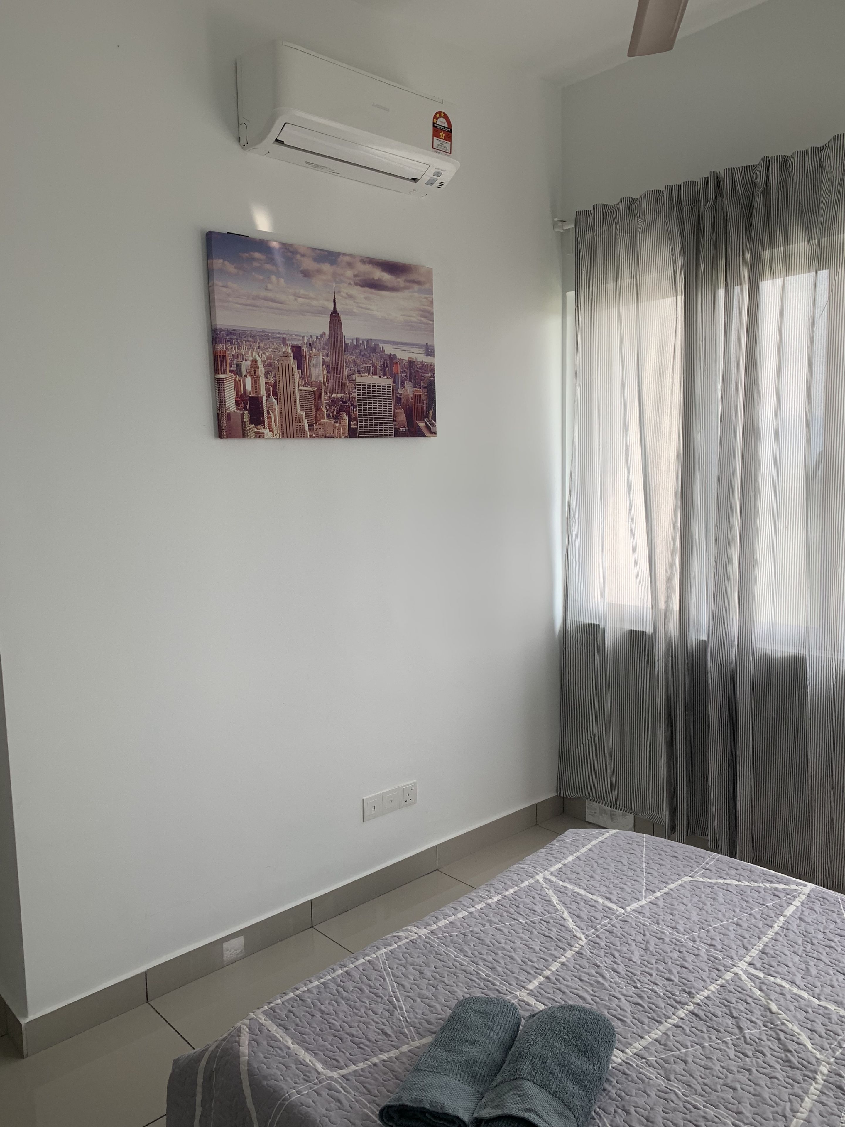 Quarto com cama, pintura de paisagem urbana, aparelho de ar condicionado e janela com cortinas.