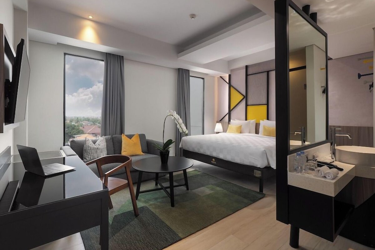 Une chambre d’hôtel moderne comprenant un lit, un bureau, une chaise, une petite table, un canapé, une grande fenêtre et une salle de bain avec un miroir et un lavabo.