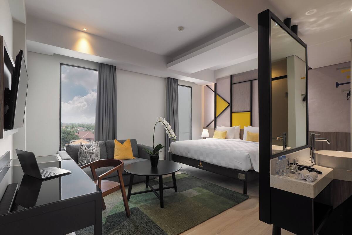 Ein modernes Hotelzimmer mit Bett, Schreibtisch, Stuhl, einem kleinen Tisch, einer Topfpflanze und einem Badezimmer mit Waschbecken und Spiegel.