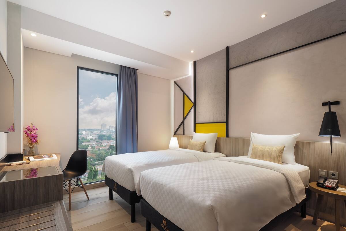 Ein modernes Hotelzimmer mit zwei Betten, einem Schreibtisch, einem Sessel und Blick auf die Stadt.