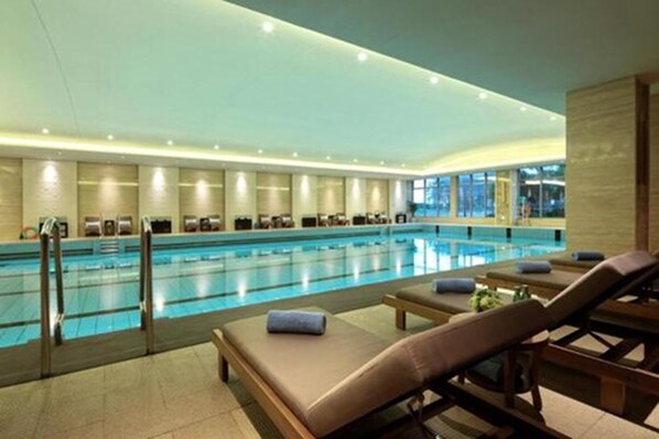 A large indoor swimming pool with multiple lanes, surrounded by lounge chairs and a glass wall.