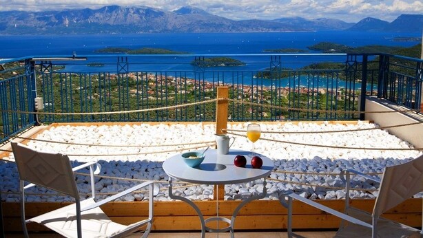 A terrace with a table, chairs, and a view of a coastal town and the sea. - Violitzis Villas (Lefkada)