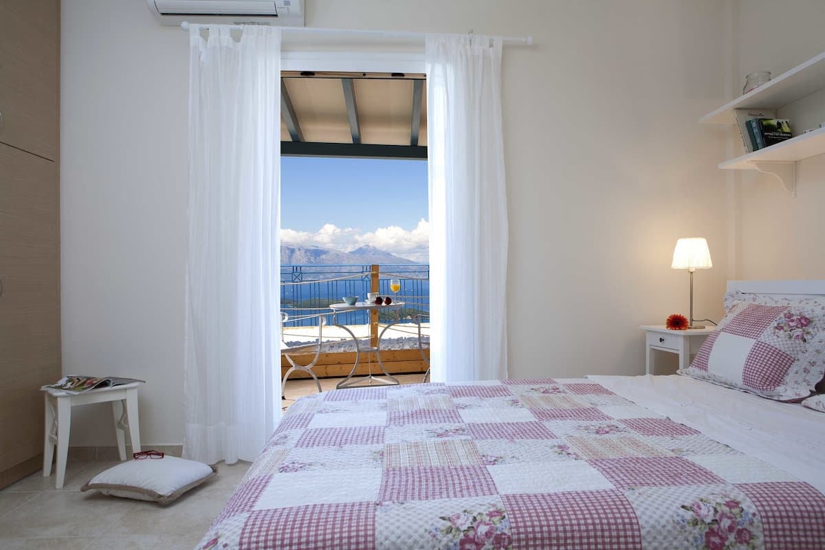 Une chambre à coucher comprenant un lit, une table de chevet, une commode, une fenêtre avec des rideaux, et offrant une vue sur la mer et les montagnes.
