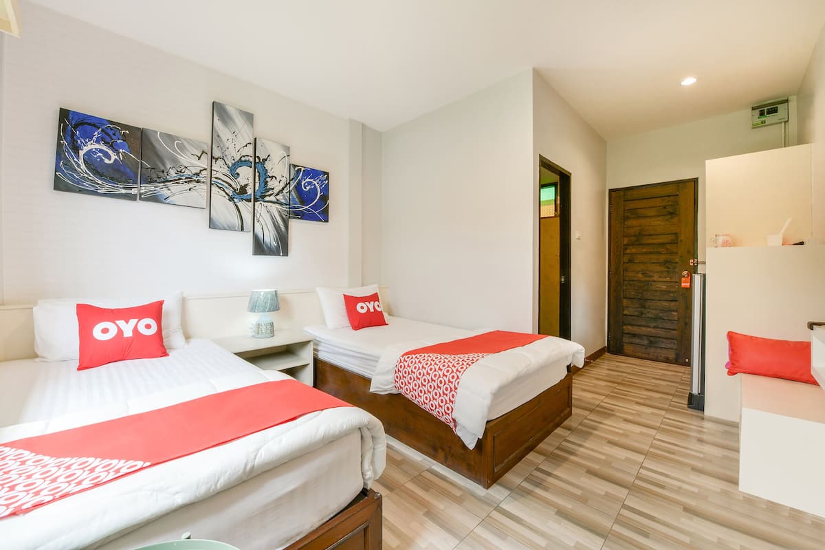 Une chambre d’hôtel avec deux lits, chacun agrémenté d’un oreiller rouge à l’effigie de la marque OYO, d’une tête de lit en bois et d’une couvre-lit blanche. Des œuvres d’art abstraites ornent les murs au-dessus des lits, et une porte en bois est visible au fond.