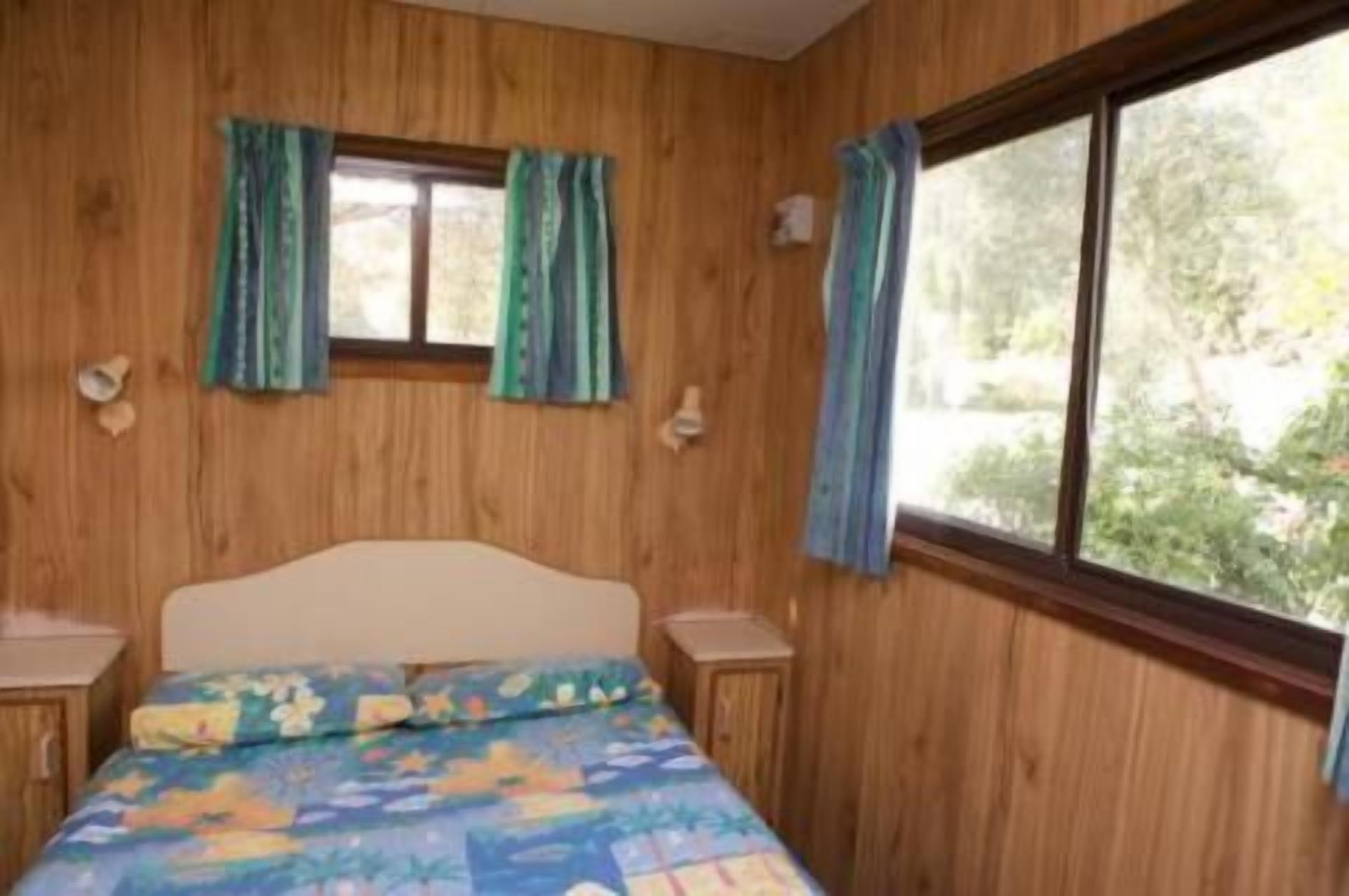 A bedroom with wooden walls, a bed with a colorful bedspread, two bedside tables, and a window with curtains.