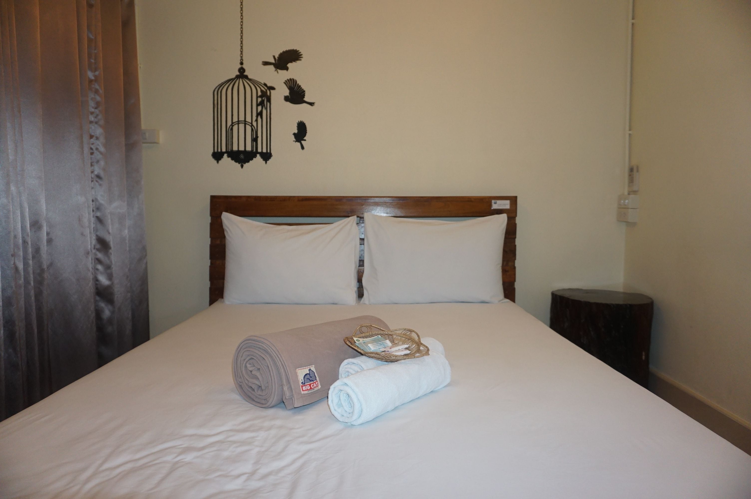 A neatly made bed with a rolled towel and a small basket, a wooden headboard, and a wall with a bird-themed decoration.