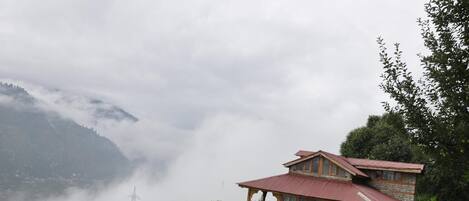 A two-story stone building with a red roof, surrounded by lush greenery and mountains in the background.