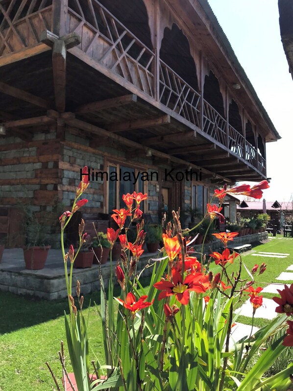 Un bâtiment traditionnel en bois, doté d’un balcon, entouré d’un jardin bien entretenu où poussent des fleurs rouges.