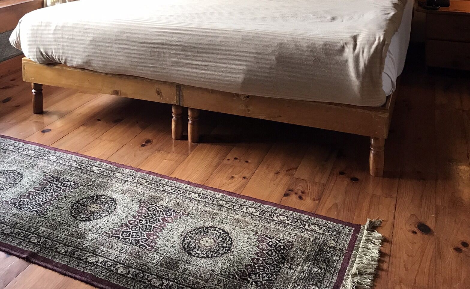 A bed with a white sheet and a wooden headboard, a patterned rug on the floor, and a wooden floor.