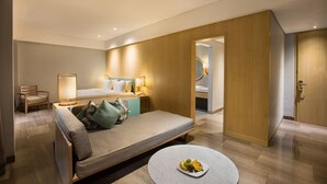Moderni hotellihuone, jossa on vuodesohva, yöpöytä lampun kanssa, pieni pyöreä pöytä, jossa on hedelmiä, ja puinen vaatekaappi.