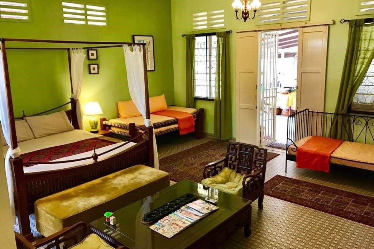 A room with a four-poster bed, a sofa, a coffee table, and a chair. There are two windows with curtains, a door, and a chandelier.