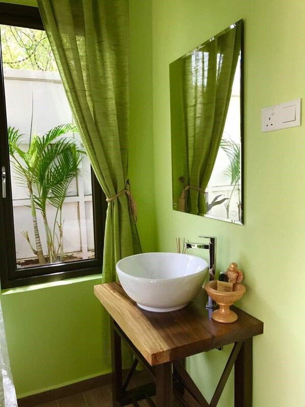 Une salle de bain avec un grand lavabo sur pied blanc, un meuble-lavabo en bois, un miroir et une fenêtre agrémentée de rideaux verts.