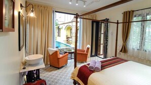 A hotel room with a bed, a sitting area with a chair and a small table, a bathroom with a sink, and a view of an outdoor pool area. - Casugria Dutch Heritage Residence (Malacca City)
