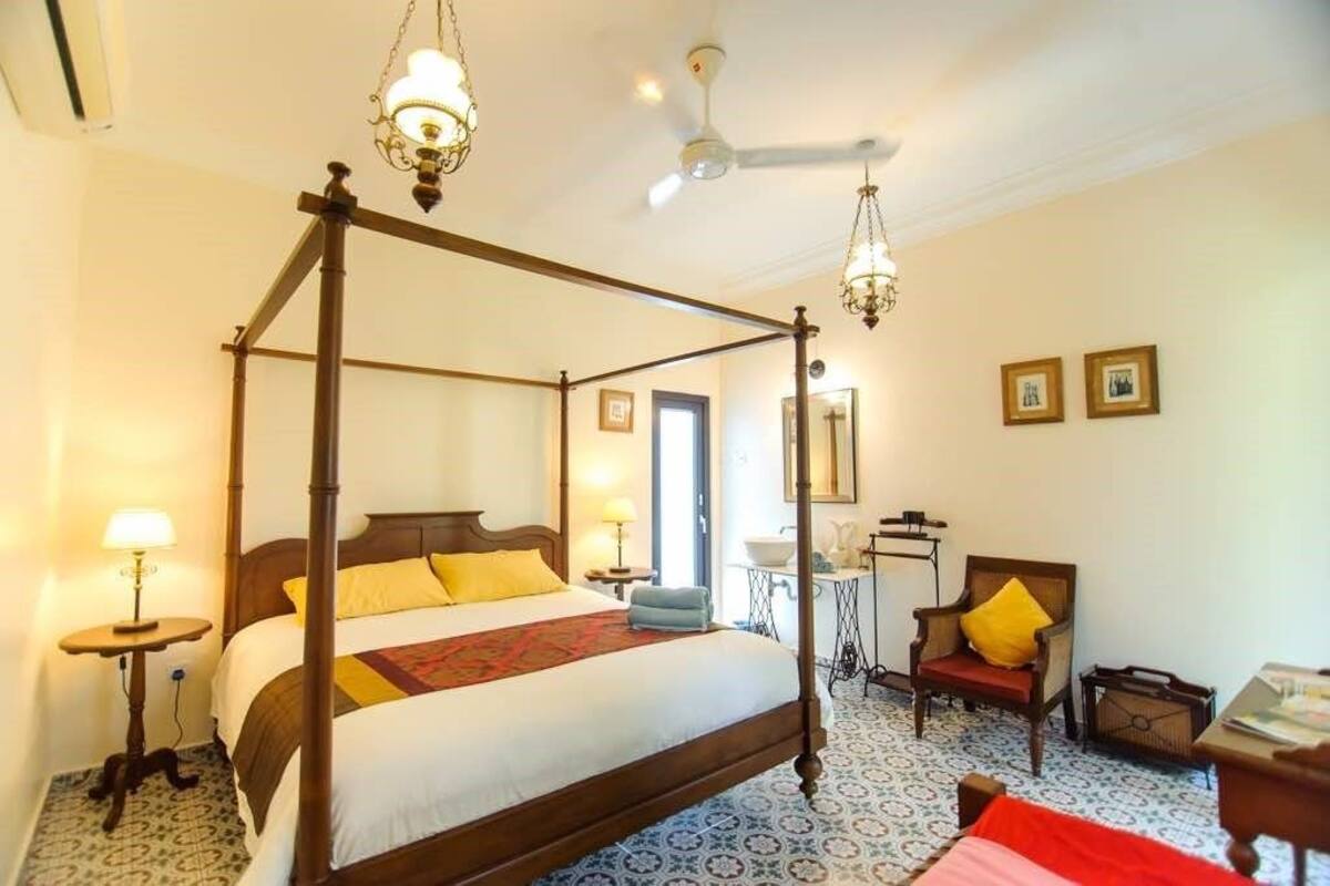 A four-poster bed with a wooden frame and a red and white bedspread. There are two bedside tables with lamps, a chair with a yellow cushion, and a small table with a mirror and decorative items.