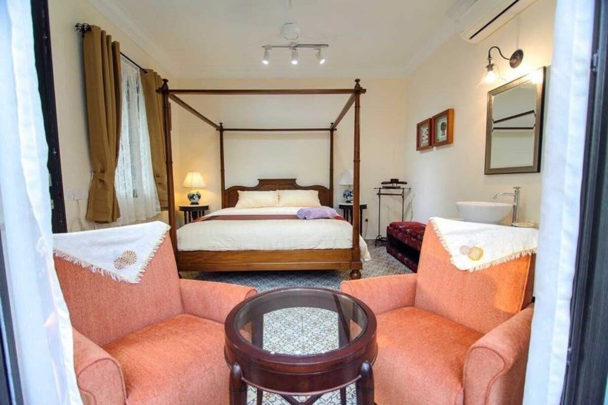 A bedroom with a four-poster bed, a round table, two armchairs, a bedside table with a lamp, a mirror, and a small cabinet.