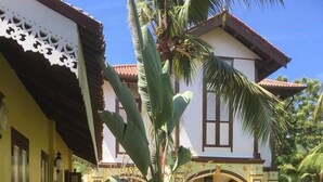 A two-story house with a yellow facade, a red-tiled roof, and a large garden with tropical plants. - Casugria Dutch Heritage Residence (Malacca City)