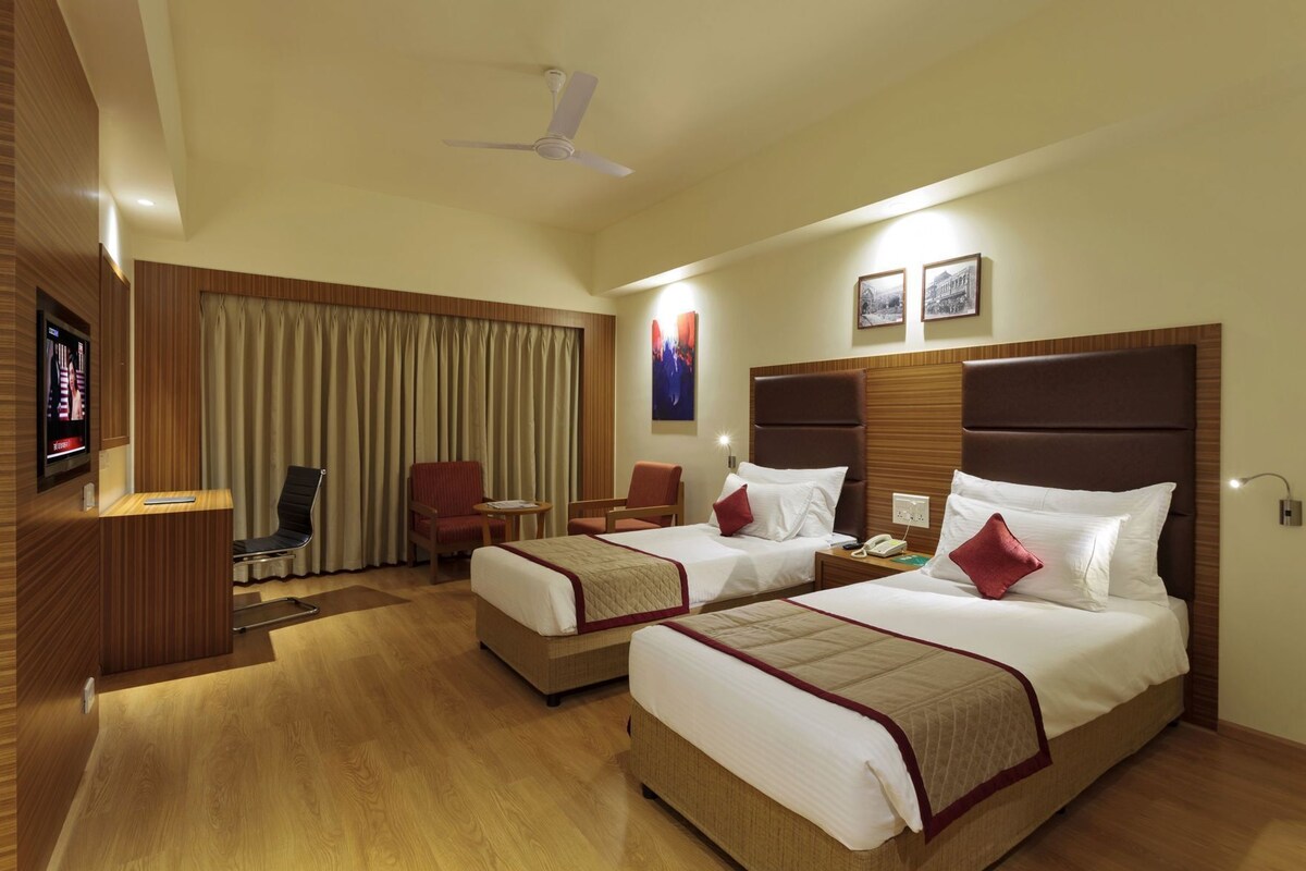 A hotel room with two beds, a desk, a chair, a TV, and a ceiling fan.