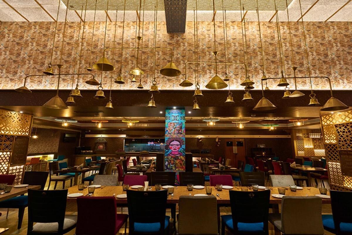 A restaurant with multiple dining tables and chairs, hanging brass chandeliers, and patterned wallpaper.
