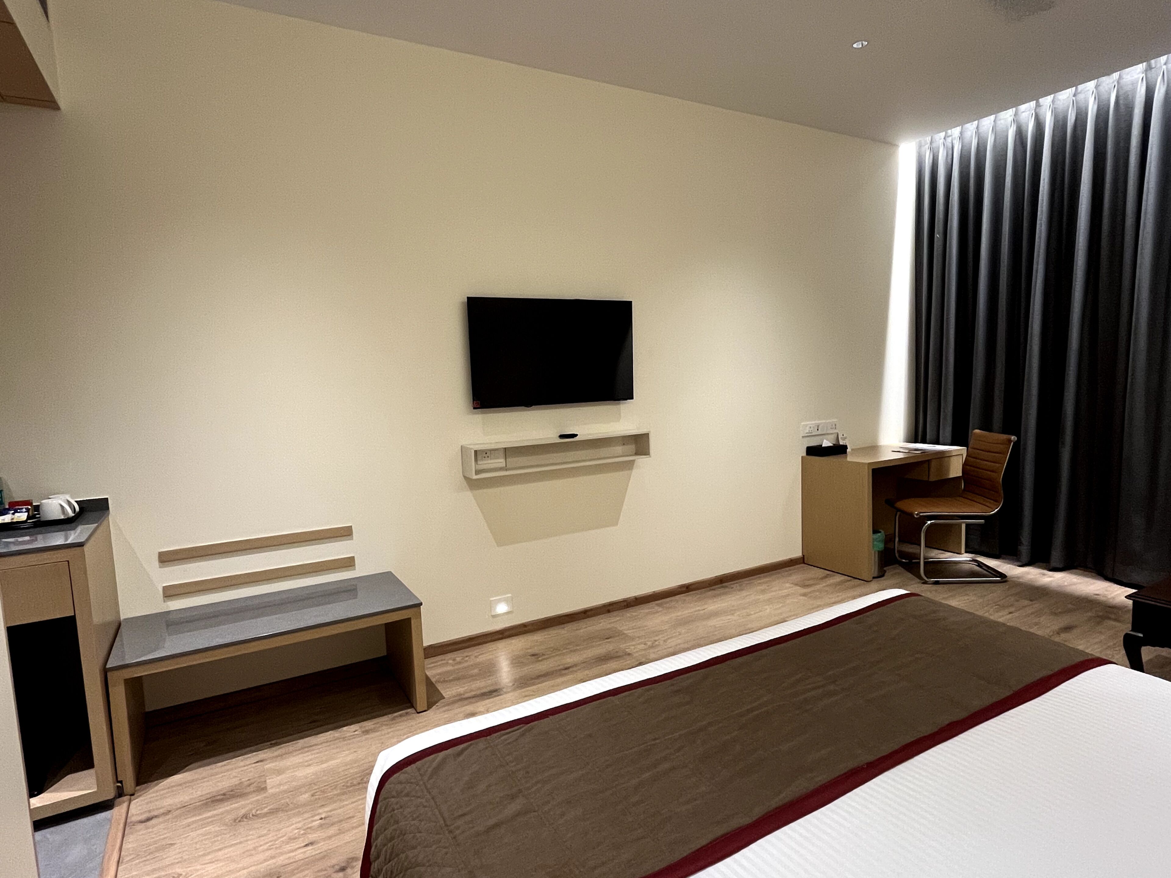 A modern hotel room with a large bed, a desk with a chair, a flat-screen TV mounted on the wall, and a window with dark curtains.