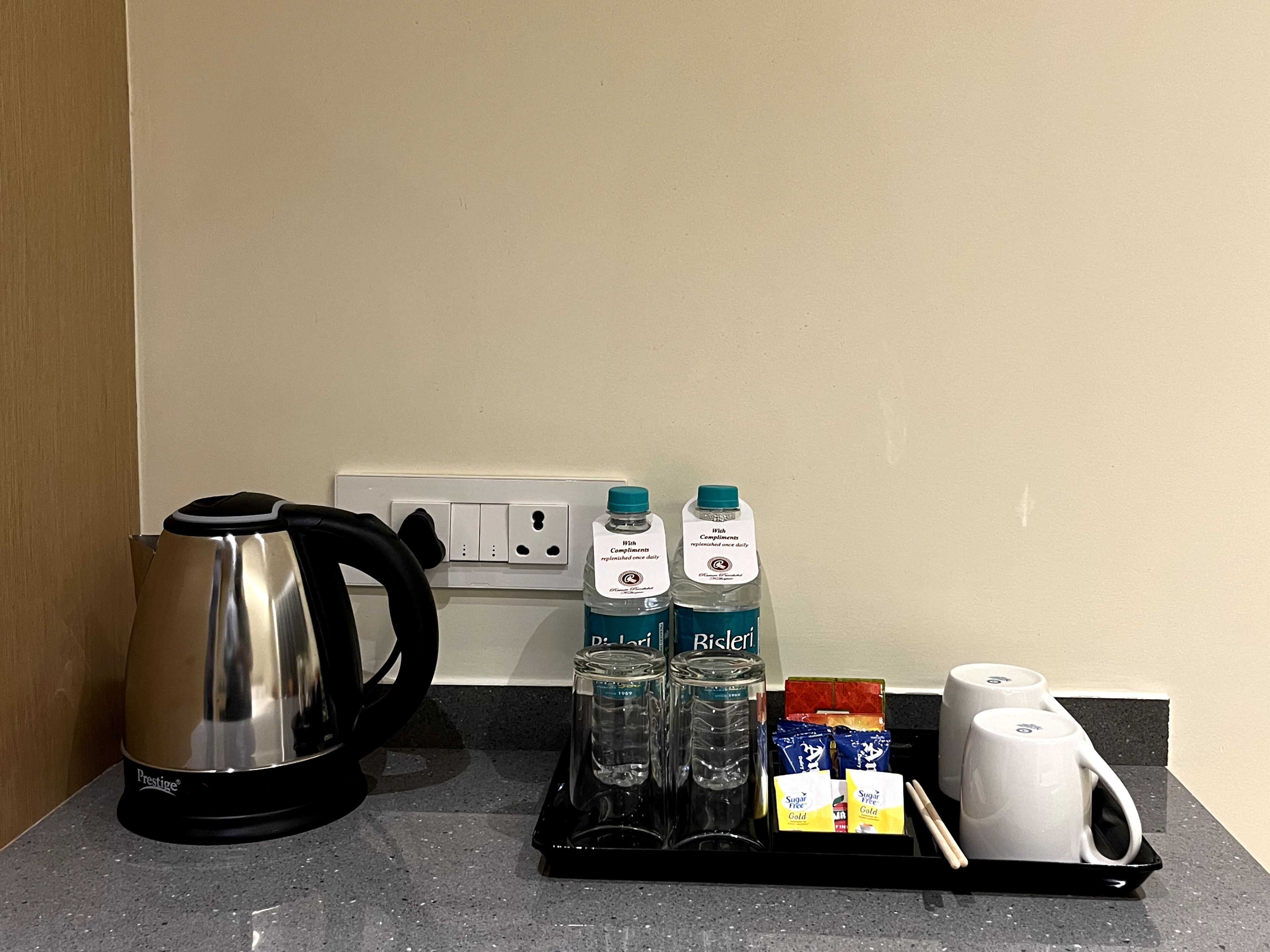 A kitchenette with a Prestige coffee maker, two bottles of water, sugar packets, and a tray with two white mugs.