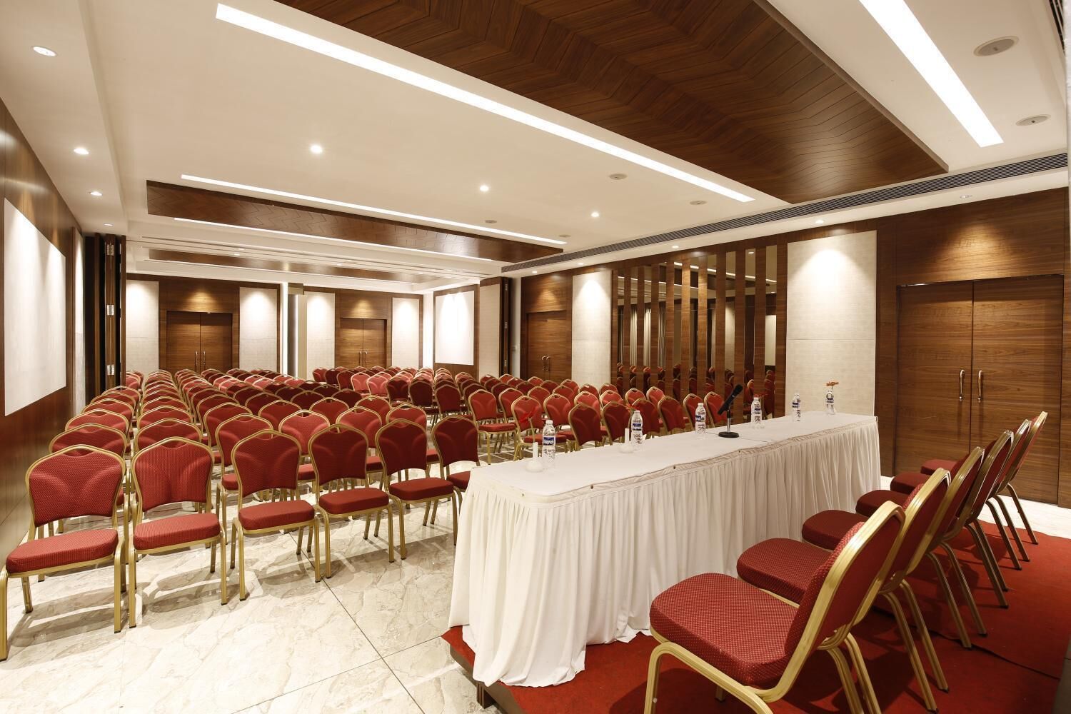 Un salón de conferencias con filas de sillas rojas, una mesa larga con mantel blanco y paneles de madera en las paredes.