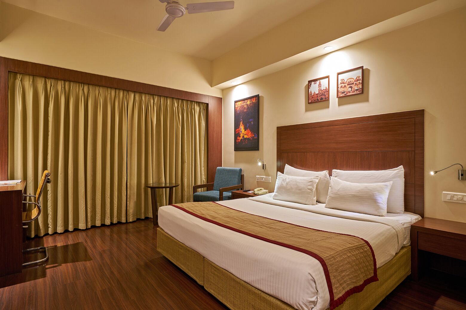 A hotel room with a large bed, wooden headboard, a desk, a chair, a small table, a telephone, and a ceiling fan.