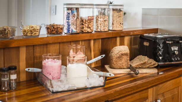 Uma bancada de cozinha com diversos alimentos e recipientes, incluindo potes de nozes e frutas secas, um liquidificador e uma barra de pão.