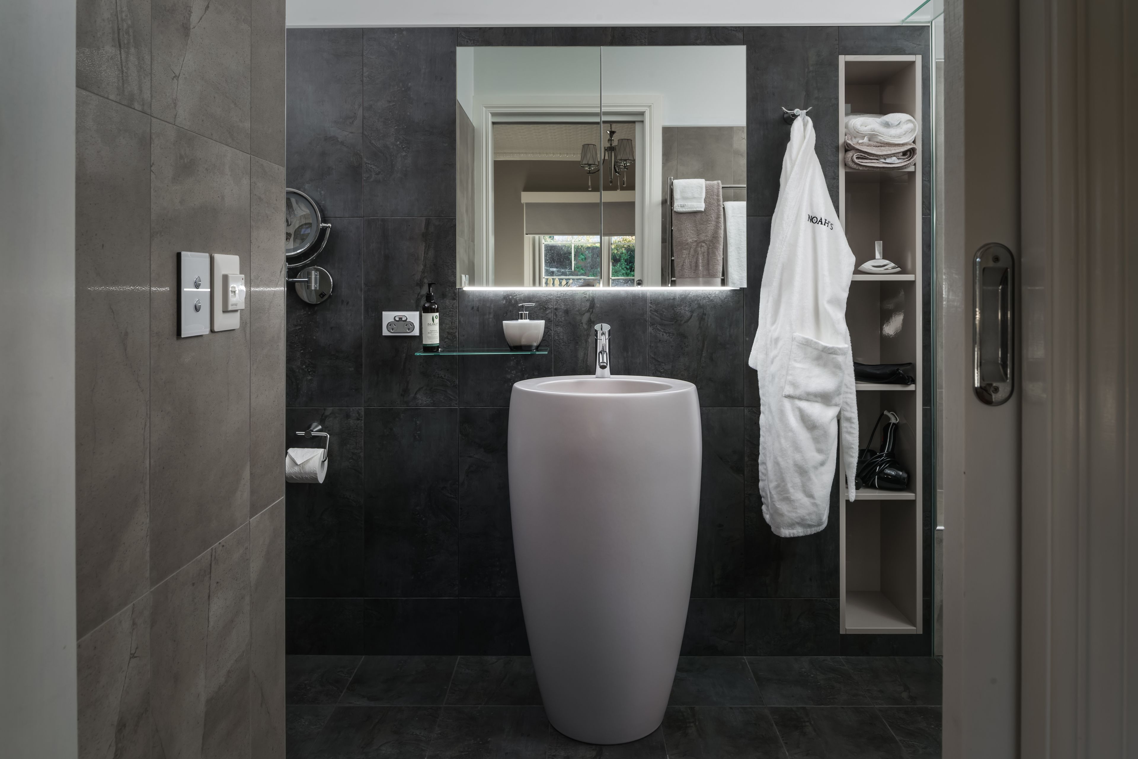 Ein modernes Badezimmer mit einem großen weißen Waschbecken, dunkel gefliesten Wänden, einem Spiegel und einem Handtuchhalter.