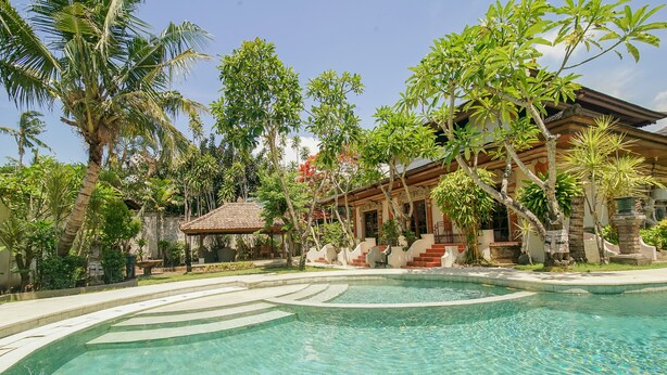 Una piscina de diseño curvo, rodeada de vegetación tropical y un edificio tradicional con tejado de baldosas.