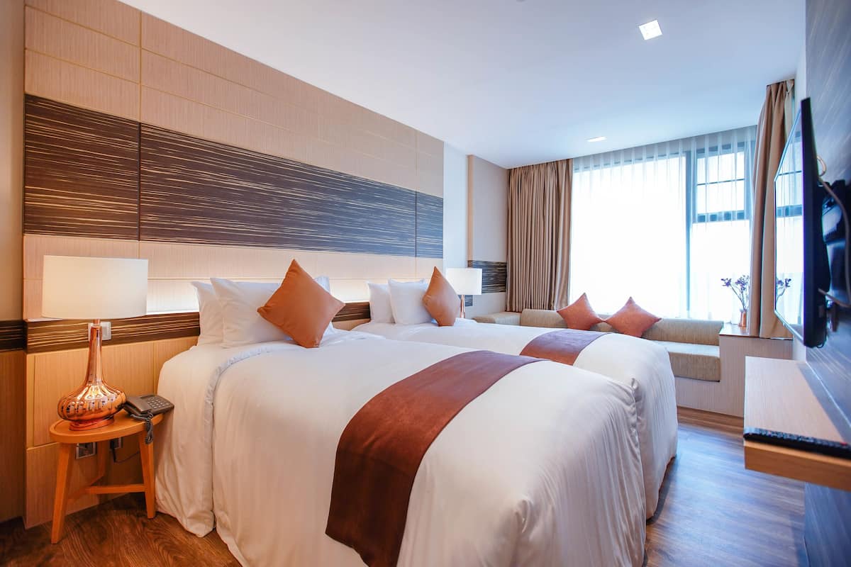 Habitación de hotel con dos camas, una mesita de noche con lámpara, televisión y un amplio ventanal con cortinas.