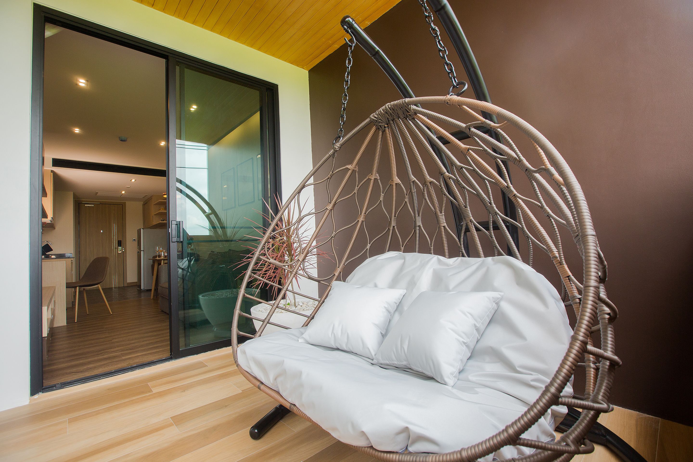 A modern interior with a wicker hanging chair with cushions, a wooden floor, and a glass door leading to a kitchen area.