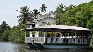 Una casa flotante con terraza cubierta y barandilla, ubicada en una masa de agua rodeada de exuberante vegetación.