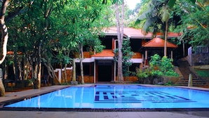 Una piscina con solárium de baldosas, rodeada de exuberante vegetación y un edificio con techo de tejas.