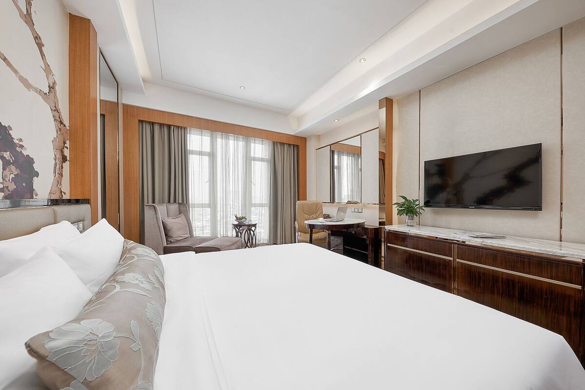 Habitación de hotel moderna con una cama grande, un escritorio, una silla, un televisor y una ventana con cortinas.