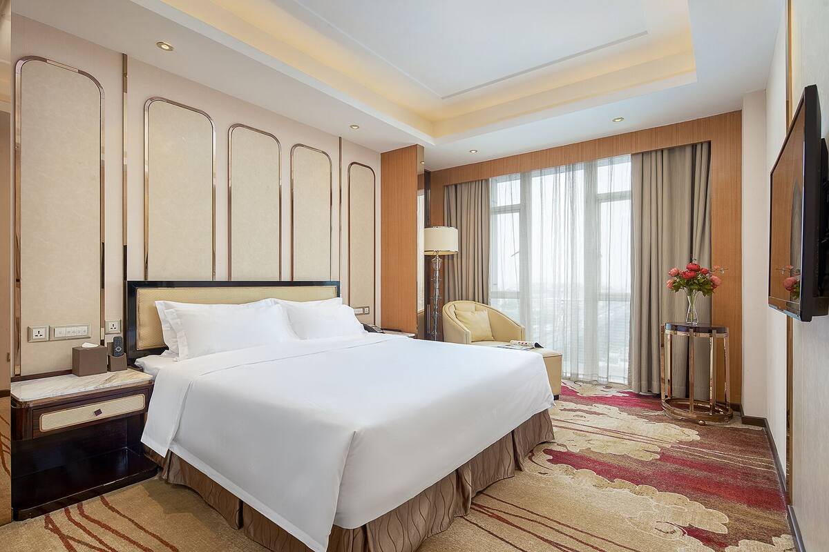 Habitación de hotel con una cama grande, una mesita de noche, una silla, un televisor y una ventana con cortinas.