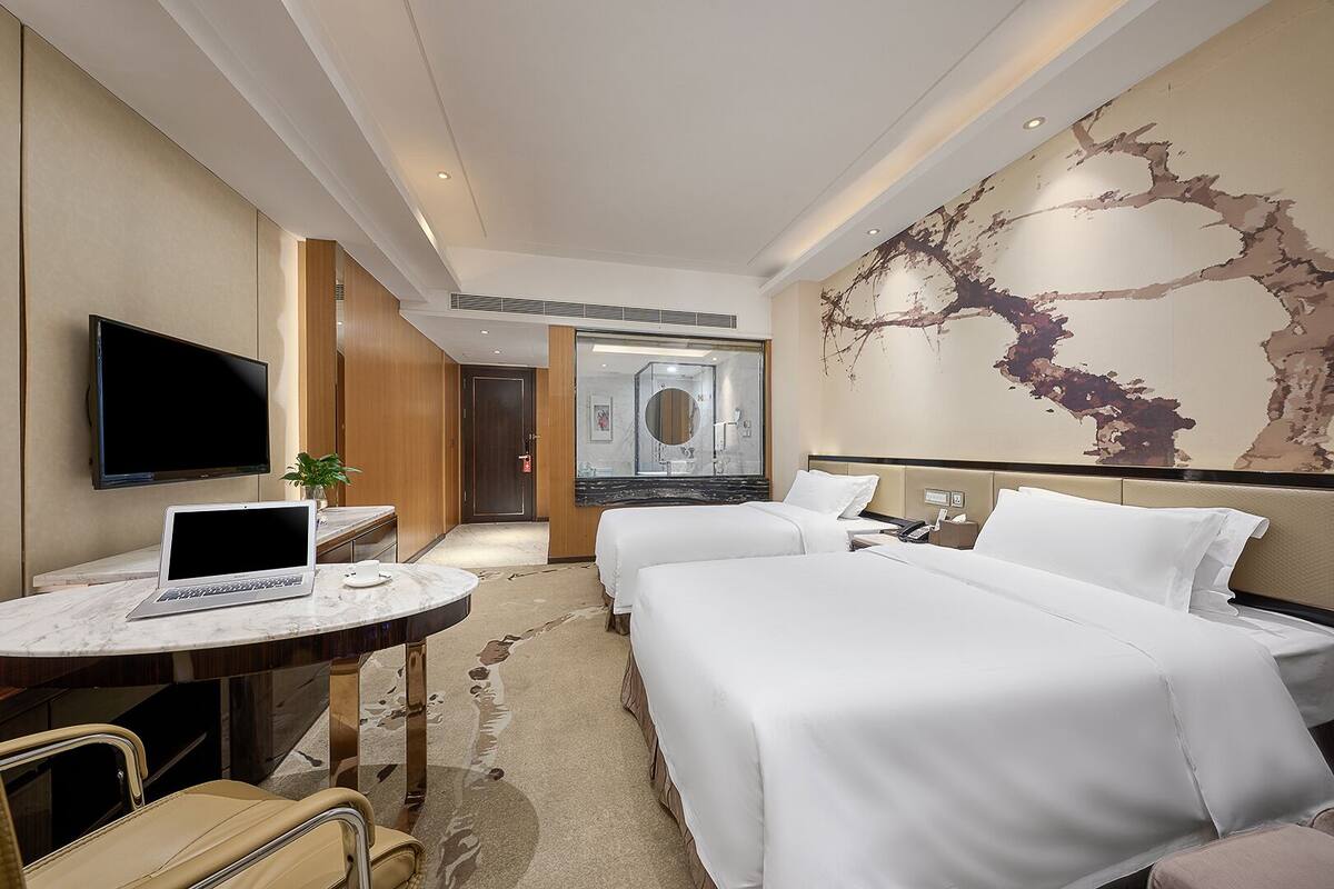 Habitación de hotel con dos camas, un escritorio con una computadora portátil, un televisor y un mural abstracto de gran tamaño en la pared.