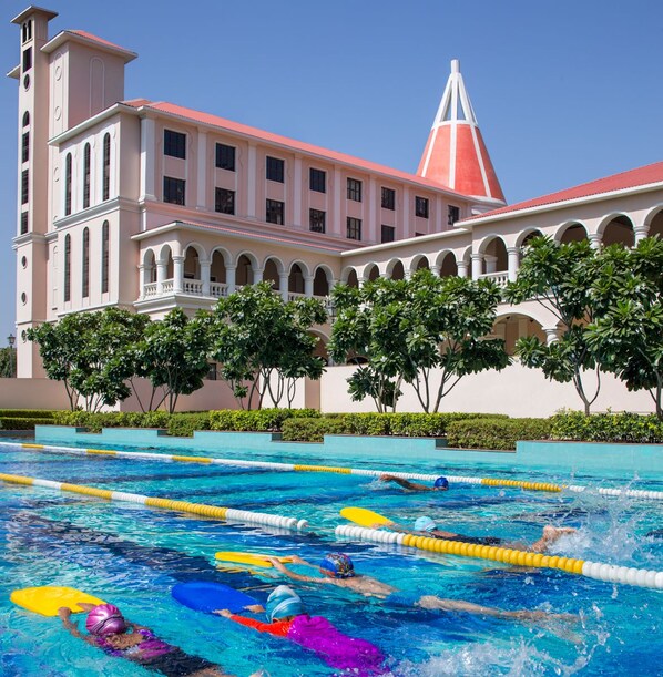 A swimming pool with lanes and swimmers, a large building with a pink roof and white columns, and a clear blue sky. - DLF Club 5 (Gurugram)